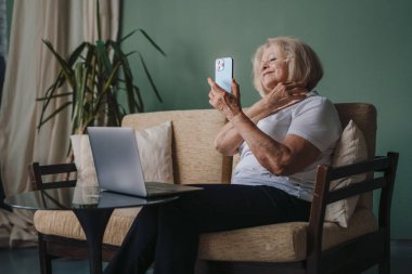 Gülümseyen gri saçlı kadın dizüstü bilgisayar kullanıyor, cep telefonuyla video görüşmesi yapıyor, evdeki rahat koltukta oturuyor. Blogger video kaydı