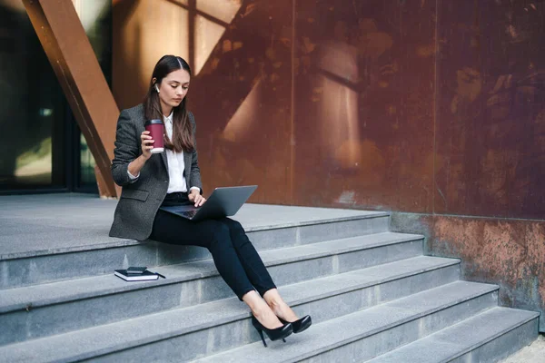 Attractive business woman drinking coffee and working with laptop while sitting on stairs outdoors. Modern communication. Attractive beautiful girl.