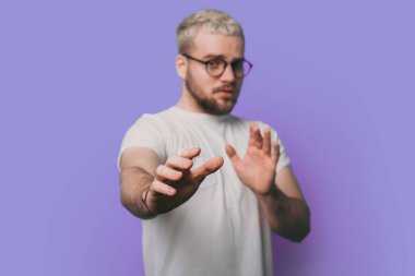 Gözlük takan beyaz bir adam tiksinti ifadesi takıyor. Çünkü tiksinti tepkisi veriyor. Elleri havada, mor arka planda izole bir şekilde duruyor..