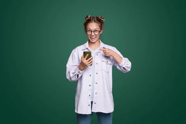 Portrait of an excited beautiful woman wearing eyeglasses pointing finger at mobile phone isolated over green background. Isolated female portrait. Beauty