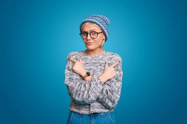 Doubting woman crossing her arms over and pointing to opposite sides, wearing eyeglasses and hat, isolated on blue background. Copy space. Choosing between two