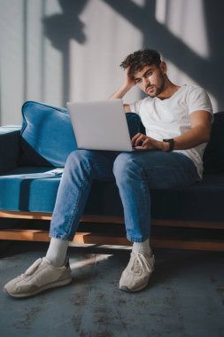 Handsome man working on laptop computer while sitting on a sofa in cozy living room. Freelancer working from home. Browsing internet. using social networks.
