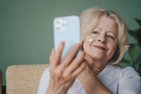 Attractive senior woman holding smart phone in front of her, using display as mirror while applying eye patches under her eyes. Smart phone. Internet