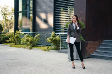 Female entrepreneur standing in front of office building with laptop and talking on phone. Walking on street, going to work.