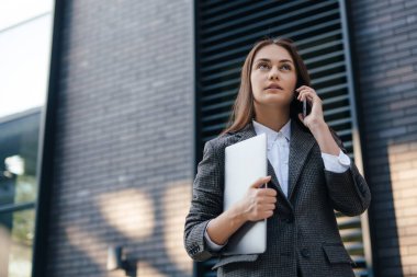 Beautiful caucasian woman talking on a mobile phone while going to the office with a laptop. Young freelancer girl is calling to the clients