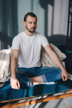 Handsome man meditating while holding remote controller of air conditioner sitting with cross-leggs on sofa. Basic yoga workout that helps to fit body and mind.