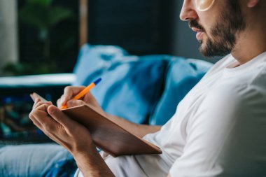 Young man with eye patches studying from home writing in a notebook with a pencil, sitting on a sofa. Sitting in living room in the evening. Student studying