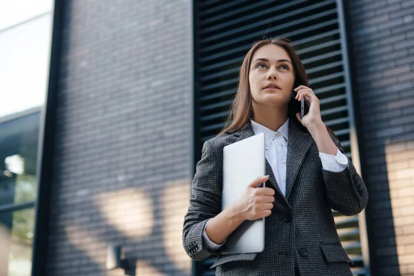 Beautiful caucasian woman talking on a mobile phone while going to the office with a laptop. Young freelancer girl is calling to the clients