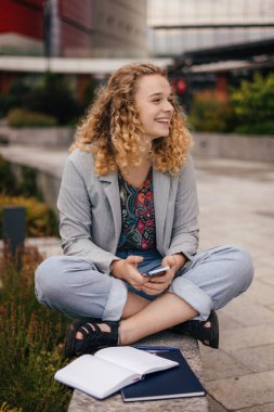 Smiling curly haired woman having opened notepad, using cellphone for surfing internet and chatting in online chat, sitting outdoors against blurred background