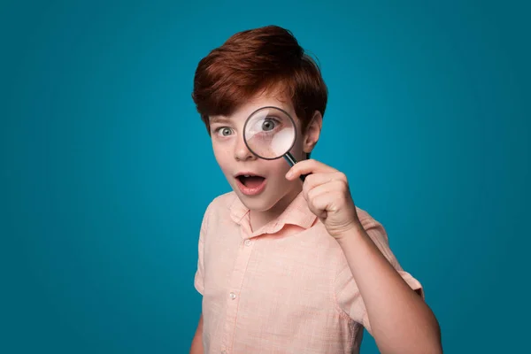 Amazed boy looking through magnifying glass, isolated on blue background. Portrait of cute little boy. Big boy eye.