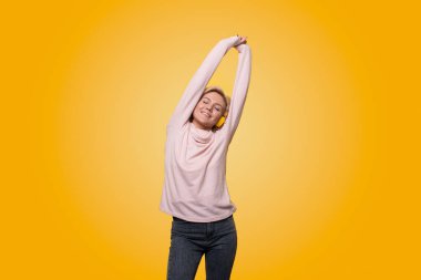 Portrait of a cheerful stylish young woman standing isolated over yellow background, listening to music with headphones, dancing