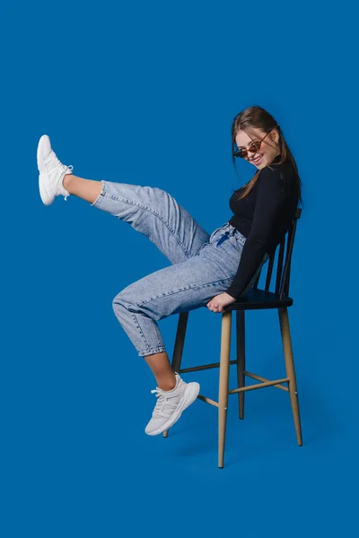 Photo of charming woman wearing eyeglasses sitting on chair rising legs isolated over blue color background.