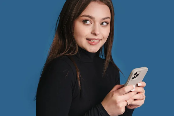 Pensive young woman biting lips holding modern cellular texting messages isolated over blue background. People technology concept