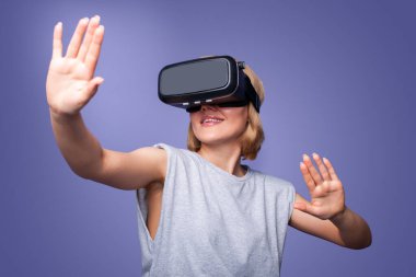 Smiling woman confidence wearing VR headset device touching air during virtual reality experience isolated over purple background. Digital technology