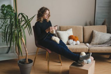 Attractive woman sitting and relaxing on comfortable chair at home on weekend enjoying her hobbies - knitting a scarf, a sweater. People lifestyle. Modern style