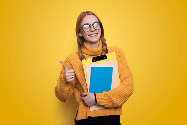 Happy teen woman with notebooks smiling and showing thumb up gesture while approving education isolated over yellow background