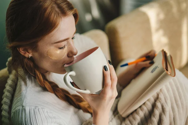 Woman with eye patches sitting on the couch holding a sketchbook in her hand and drinking a cup of coffee. Rest, relaxation, education, training, home comfort.