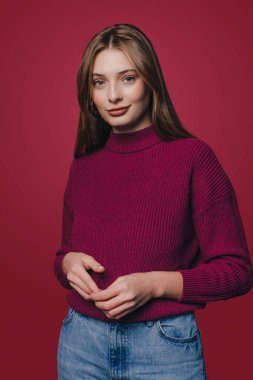 Portrait of confident beautiful woman wearing casual clothes, standing in relaxed pose, smiling and looking at camera, isolated over crimson studio background