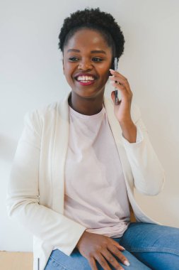 Young attractive African woman talking on cell phone to her friend, looking cheerful, joyful and happy, sitting at the window. Human emotions and feelings