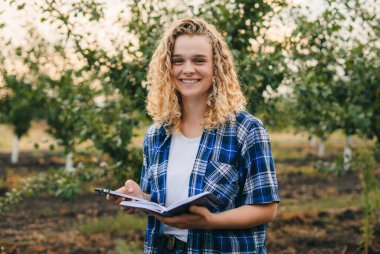Curly haired farmer woman checking plum orchards progress, noting in her textbook. Harvesting. Agro business.
