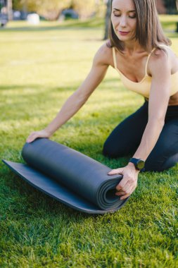 Young woman sitting on the grass and rolling yoga mat finishing outdoor workout in the morning in the park. Happy lifestyle. Fitness woman.