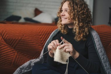 Curly haired young woman wrapped in a blaket holding knitting needles sitting on the floor in living room and enjoying looking away while smiling with teeth
