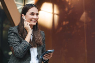 Close up portrait of brunette businesswoman with wireless headphones standing outside the office building holding in hand mobile phone chatting with friends
