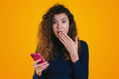 Photo of shocked woman blogger watching video using mobile phone, looking with big eyes, covering mouth isolated on yellow wall background