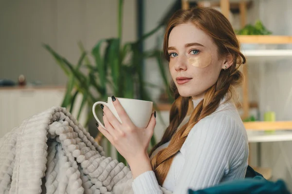 Relaxed young redhead woman enjoying a tea break sitting wrapped in a warm blanket on a comfortable sofa looking at camera. Domestic lifestyle. Happy lifestyle.
