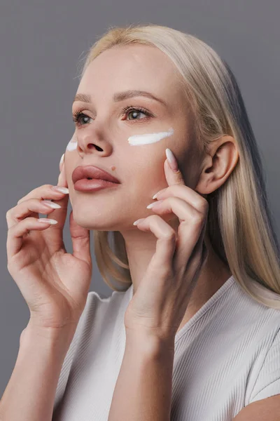 Closeup of woman applying anti-aging or moisturising face cream on gray studio background. Face care, lifting, anti-aging, nourishing cosmetics concept
