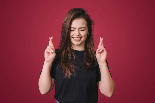 Pleased cheerful brunette woman wishing good luck, crossing fingers, having faith for better, isolated over crimson background. People, body language concept