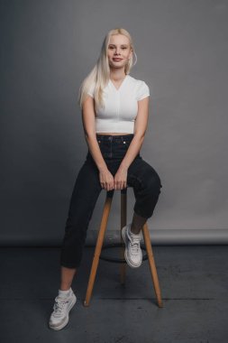 Blonde caucasian attractive woman model posing in studio wearing white t-shirt sitting on chair, isolated on gray studio wall background