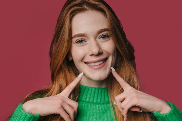 Read headed woman with freckles on her face standing over crimson background smiling cheerful showing and pointing with fingers teeth and mouth. Dental health