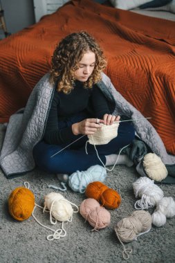 Scene portrait of curly-haired young woman doing knit work at home sitting on the floor wrapped in a blanket. People and hobby. Indoor leisure activity female