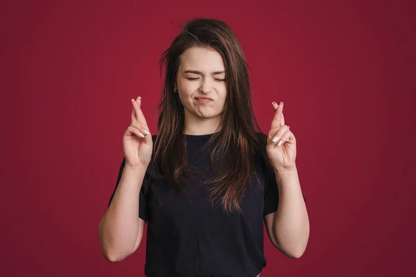 Hopeful brunette woman crossing fingers for good luck being dressed in a black t-shirt closing eyes isolated over red background. Female student making wish
