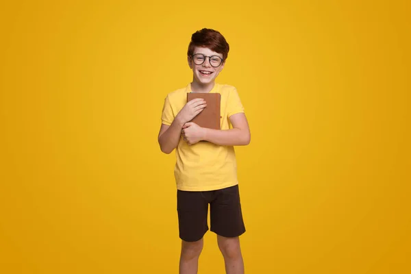 Adorable little boy with ginger hair wearing round eyeglasses and classy outfit, holding book and looking at camera against yellow background. Information