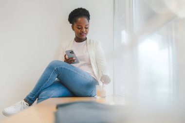 An afro woman holding and using smart phone while drinking iced coffee at coffee shop. Smart device. Business lifestyle. Smart phone.
