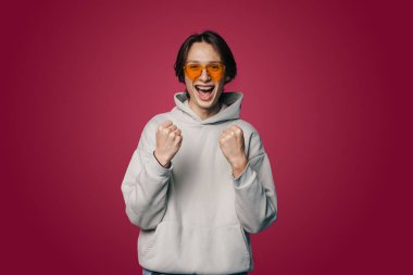Happy joyful young man wearing eyeglasses doing winner gesture clenching fists isolated on pink color wall background studio portrait