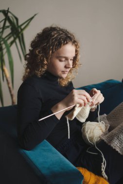 Young woman sitting on sofa at home holding knitting needles and yarn knits clothes. Favorite activity and pastime. Retired tranquil carefree life concept