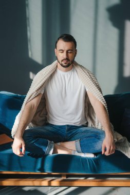 Calm relaxed caucasian man, sitting alone at home in living room on a sofa, meditating in the lotus position, take care of his mental health. Chilling and