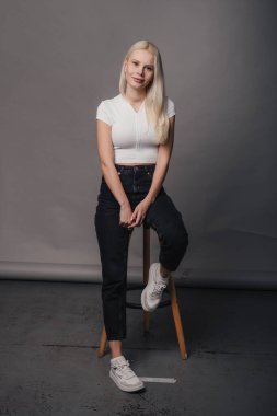 Full length portrait of a smiling casual blonde woman sitting on a stool and looking at camera posing over gray background. Happy lifestyle. Attractive