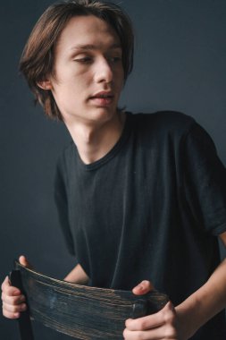 Pensive brunette man in black t-shirt looking away while sitting on the chair posing over black background. Confident man. Fashionable style. Young male model