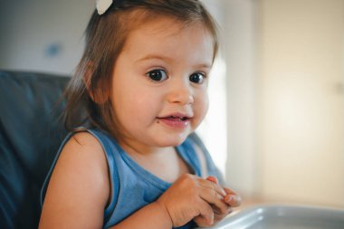 A small baby girl looking at camera while sitting on a highchair and eating her meal. Healthy food. Healthy eating.