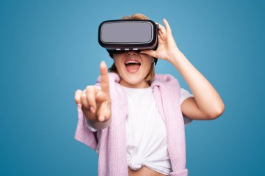 Cheerful young woman using VR glasses playing video games online, using modern gadgets touching copy space isolated on blue background. Cyberspace concept