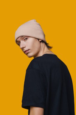 Portrait of a confident young man dressed in a black t-shirt wearing hat looking over his shoulder at camera isolated on yellow background. Studio fashion