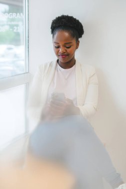 Smiling female student looking down at phone screen and making notes sitting at cafe shop. African american girl sitting at cafe with mobile phone. Black woman