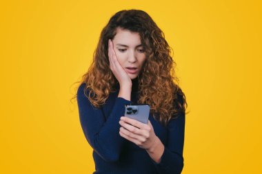 Portrait of a surprised confused woman with long curly hair looking at mobile phone isolated over yellow background. Text message. Holding smartphone. Wow