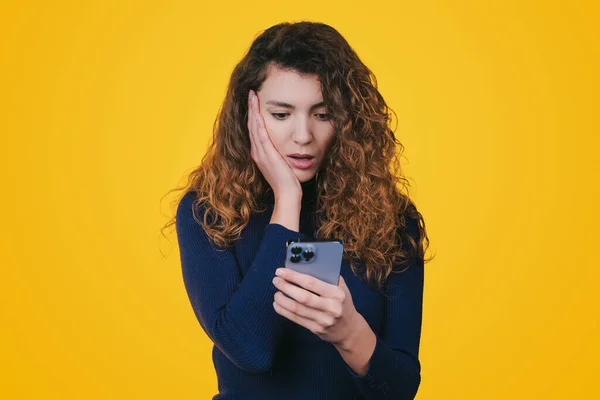 Portrait of a surprised confused woman with long curly hair looking at mobile phone isolated over yellow background. Text message. Holding smartphone. Wow