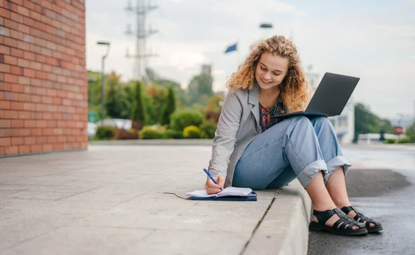 Portrait of young woman student making notes in notepad while sitting outdoors with laptop computer. The Lifestyle of university students outside the home