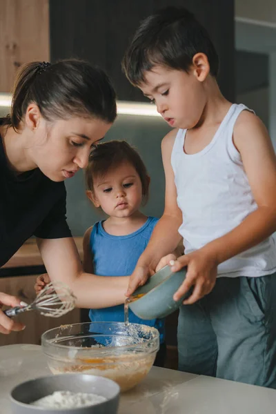 Caucasian mother having fun baking in modern kitchen with her little kids, prepare biscuits, pancakes for breakfast at home. Funny weekend. Baking together